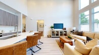 This two-story home is designed with a bright, open-concept layout that seamlessly connects the main living areas. The living room is a standout feature, boasting dramatically high ceilings and a wall of large windows that flood the space with natural light and provide views of surrounding greenery. Light-toned hardwood-style flooring runs throughout the main level, contributing to the home's clean, contemporary aesthetic.

The kitchen is well-appointed with flat-front cabinetry in a warm gray wood-grain finish, stainless steel appliances including a range and microwave, and a white quartz-topped island with bar seating for casual dining. Adjacent to the kitchen, a dedicated dining area features a stylish geometric chandelier and access to natural light through large vertical blinds.

A half bathroom on the main floor adds convenience for guests. A dedicated office space, finished with plush carpet, provides a quiet and functional work area. The in-unit laundry room houses a full-sized washer and dryer.

Upstairs, the carpeted bedroom offers a comfortable, well-lit retreat with ample natural light. A secondary lounge or loft area provides additional flexible living space. Throughout, the home maintains a neutral, cohesive palette that creates a calm and inviting atmosphere.