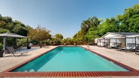 Located at 9989 Burbank Dr in Baton Rouge, Louisiana, this outdoor pool area offers a well-appointed recreational retreat designed for relaxation and leisure. The centerpiece is a generously sized rectangular swimming pool with clear turquoise water, framed by classic brick coping and a broad concrete deck that provides ample space for lounging. Depth markers are visible along the pool's edge, and safety equipment including a life ring is mounted near the perimeter fence, reflecting a commitment to guest safety.

The deck is furnished with multiple lounge chairs and seating arrangements, catering to both sunbathing and social gatherings. A standout feature is the freestanding gazebo with a dark canopy, positioned poolside and surrounded by additional outdoor seating and decorative potted plants, offering a shaded retreat on sunny Louisiana days. Ornamental lamp posts dot the perimeter, providing ambient lighting for evening use.

The entire area is enclosed by a sturdy wrought iron fence with a gated entry, which also displays posted pool rules and regulations. Mature trees and manicured landscaping frame the space on all sides, creating a sense of natural privacy and a cool, verdant backdrop. The open sky above and surrounding greenery lend the area a bright, welcoming ambiance throughout the day.