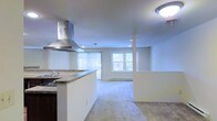 This single-floor residence offers a clean, open layout that seamlessly connects the kitchen and living areas into one cohesive, functional space. The kitchen is outfitted with a striking stainless steel wall-mounted range hood as a standout feature, complemented by a granite-topped peninsula that provides both prep space and a natural divider between cooking and living zones. A double sink and dark cabinetry add contrast to the light-toned walls and neutral finishes throughout.

The open-plan living area extends generously from the kitchen, providing ample room for a variety of furniture arrangements. Soft, neutral carpeting covers the living space floor, creating a warm and comfortable atmosphere. Recessed lighting and a ceiling-mounted light fixture ensure the interior remains well-lit, while multiple windows dressed with vertical blinds invite natural light deep into the room. One window offers a pleasant view of greenery outdoors, enhancing the sense of connection to the outside. A baseboard heating unit runs along the exterior wall, ensuring year-round comfort. The bright white walls and open sightlines contribute to an airy, welcoming ambiance throughout the entire space.