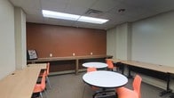 Located at 364 W Lane Ave in Columbus, Ohio, this single-floor space is designed to support collaborative work, study, and casual gatherings. The room features a practical and organized layout, anchored by a series of round white tables paired with vibrant orange chairs that encourage group interaction and discussion. Along one wall, a continuous built-in wooden counter provides additional workspace, ideal for individual tasks or laptop use, with conveniently placed electrical outlets for easy access to power. Rectangular tables with matching seating are also available, offering versatile arrangements to accommodate a variety of needs. The space is finished with a drop ceiling fitted with recessed fluorescent panel lighting that ensures consistent, well-distributed illumination throughout. A bold terracotta-toned accent wall adds a warm, energetic character to the interior, complementing the neutral carpeted flooring. The overall design balances functionality with a welcoming ambiance, making it well-suited as a study lounge, meeting room, or multipurpose gathering area. The straightforward, open layout allows for easy reconfiguration to suit different group sizes and activities.
