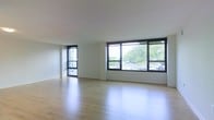 This single-floor space offers a clean and modern aesthetic defined by an open, airy layout that feels both expansive and welcoming. The interior is anchored by large, dark-framed windows that span a significant portion of the exterior wall, flooding the space with abundant natural light and providing views of lush greenery and the surrounding streetscape beyond. The windows are generously proportioned, creating a seamless visual connection between the interior and the outdoors. The flooring throughout is a light-toned hardwood, lending warmth and continuity to the space while reflecting natural light to enhance the overall brightness. The walls are finished in a crisp, clean white, contributing to a minimalist atmosphere that feels fresh and versatile. The smooth white ceiling complements the neutral palette and adds to the sense of openness. The overall ambiance is calm and uncluttered, making the space well-suited for a variety of uses. The combination of expansive glazing, natural wood flooring, and understated finishes creates an inviting environment that is both functional and visually refined.