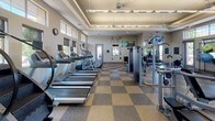 Located at Denver West Circle in Lakewood, Colorado, this single-floor fitness center is a comprehensively outfitted facility designed to support a wide range of workout routines. The space features a distinctive checkered rubber flooring in neutral tones, providing both durability and a clean, professional aesthetic throughout.

The cardio zone is well-stocked with a row of treadmills, elliptical trainers, and stair climbers, each equipped with individual screens for entertainment during workouts. Multiple ceiling-mounted televisions are positioned throughout the facility, ensuring visibility from virtually every area of the floor.

The strength training section includes a robust selection of cable machines, selectorized weight equipment, and multi-functional cable stations. A dedicated free weight area houses a full rack of dumbbells alongside adjustable benches, offering ample space for both beginner and advanced lifters.

A functional training corner is outfitted with stability balls, step platforms, and suspension training equipment, catering to flexibility and bodyweight-focused workouts. Large windows positioned along the upper walls invite abundant natural light, creating an open and energizing atmosphere while offering views of the surrounding greenery. The elevated, coffered ceiling further enhances the sense of spaciousness. The facility is well-lit with a combination of recessed and linear overhead lighting, ensuring a bright and welcoming environment throughout all hours of operation.