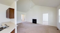 This single-story space is defined by its striking vaulted ceiling, which soars to an impressive height in the main living area, creating a sense of openness and grandeur. A ceiling fan is centrally mounted at the peak, providing both functional air circulation and a clean architectural accent against the all-white ceiling and walls.

The living area is anchored by a traditional fireplace with a white surround and black firebox, serving as a natural focal point within the room. Plush, neutral-toned carpeting extends across the floor, contributing to a warm and comfortable atmosphere throughout the space.

The crisp white walls are consistent throughout, lending the interior a bright, clean aesthetic that feels both airy and versatile. Dark wood cabinetry is partially visible along one edge of the space, suggesting an adjacent kitchen or built-in storage area that complements the neutral palette with contrast and warmth.

Multiple doorways are visible, indicating connections to additional rooms beyond the main living area. The open layout, generous ceiling height, and understated finishes combine to give this space a welcoming and functional character well-suited for a variety of uses.