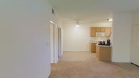 This single-floor residence showcases a practical and open layout that seamlessly connects the living area to the kitchen, creating a spacious and cohesive environment. The living area is finished with wall-to-wall carpeting in a neutral tone, contributing to a warm and comfortable atmosphere. A ceiling fan is centrally positioned overhead, providing both functionality and a subtle design element to the space.

The kitchen is situated in an open, peninsula-style configuration, featuring medium-toned wood cabinetry that extends to the upper portion of the walls. The kitchen is equipped with a gas range, a built-in over-the-range microwave, and generous counter space, making it well-suited for everyday cooking needs. The peninsula provides an informal dining or prep area that reinforces the open-concept feel.

The walls throughout are finished in a clean, crisp white, lending the space a bright and airy quality. Doorways visible from the main living area suggest access to additional rooms, including what appear to be closets or private spaces. HVAC venting integrated into the ceiling ensures year-round climate comfort. Overall, this residence offers a straightforward, functional layout ideal for comfortable everyday living.