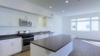 This single-floor space showcases a clean, contemporary design centered around an open-concept kitchen and living area. The kitchen is equipped with stainless steel appliances, including a gas range, built-in microwave, and dishwasher. Crisp white cabinetry lines the walls, complemented by dark quartz countertops that extend across both the perimeter workspace and a generously sized central island, providing ample prep and dining space. Recessed lighting throughout the kitchen ensures consistent illumination, while a stainless steel sink is conveniently positioned along the back counter. The open layout flows seamlessly into the adjacent living area, which benefits from a large window that invites natural light deep into the space, creating a bright and airy atmosphere. Light wood-tone flooring runs throughout, adding warmth to the otherwise neutral white palette. The minimalist aesthetic, defined by clean lines and a monochromatic color scheme, lends the space a modern, functional character well-suited for everyday living and entertaining.
