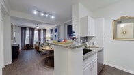 This well-appointed single-floor residence combines modern design with functional living across a thoughtfully laid-out floor plan. The open-concept kitchen features white shaker cabinetry, granite countertops, a peninsula with a built-in sink, grey subway tile backsplash, and stainless steel appliances including a range, microwave, and refrigerator. Track lighting illuminates the space, which flows seamlessly into the dining and living areas. The dining area offers built-in shelving and is open to the living room, which is anchored by a bold blue sofa, abstract artwork, and dark hardwood-style flooring that runs throughout the unit. Large windows dressed with floor-length grey curtains and wood blinds invite abundant natural light and views of the surrounding exterior. The primary bedroom is generously sized with oversized windows, a ceiling fan, and ample space for a sitting area. A second bedroom provides a cozy, well-lit retreat. The full bathroom is elongated and elegantly finished, featuring a dual-sink vanity with granite countertops, grey cabinetry, a tub-shower combination with grey tile surround, and coordinating hardwood-style flooring. Overall, the space presents a cohesive, contemporary aesthetic with quality finishes and a bright, inviting ambiance.