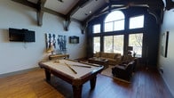 This single-story recreation and lounge space combines entertainment and relaxation in an open, generously scaled layout. The room's most striking architectural feature is its dramatic vaulted ceiling, accented by dark wood exposed beams and decorative corbels that add a refined, craftsman-inspired character. A large arched window flanked by tall rectangular panes dominates one wall, flooding the interior with abundant natural light and offering views of the surrounding landscape.

The hardwood flooring runs throughout, grounding the space with warmth and continuity. A full-size billiards table with a cream felt surface serves as the focal point of the room, positioned centrally for easy play. Adjacent to it, a comfortable lounge area features a neutral-toned sofa, armchairs, and a wooden coffee table arranged on a textured area rug, creating an inviting space to relax.

A wall-mounted flat-screen television is positioned above a decorative console, accompanied by a table lamp and bold abstract wall art. Recessed lighting supplements the natural light. The overall ambiance is upscale yet approachable, blending rustic architectural details with modern amenities for a well-rounded entertainment environment.
