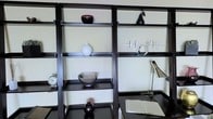 This single-floor space showcases a refined modern aesthetic defined by clean lines and thoughtful design. The focal point of the room is a striking built-in shelving unit crafted in a deep, dark-toned finish, offering multiple tiers of display space. The shelves are adorned with a carefully curated collection of decorative objects, including ceramic vases, sculptural figurines, a classic analog clock, and contemporary metallic accents, all contributing to an atmosphere of understated elegance. The warm, neutral wall tones provide a soft contrast to the bold shelving structure, enhancing the visual depth of the space. Integrated seamlessly into the shelving unit is a sleek desk surface, complete with an adjustable task lamp and an open book, suggesting a functional workspace that blends harmoniously with the decorative elements. The overall ambiance is sophisticated and intentional, balancing form and function with a consistent monochromatic color palette of blacks, whites, and silvers. This space is ideal for those who appreciate a contemporary interior where every detail is purposefully placed.