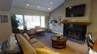 This single-floor space is designed with comfort and style in mind, centered around an inviting living area that immediately draws the eye. The vaulted ceiling adds a sense of openness and grandeur, while track lighting provides flexible, modern illumination throughout the room. A striking dark-tile fireplace serves as a natural focal point along one wall, topped with a warm wood mantel and a wall-mounted flat-screen television above, creating an ideal gathering spot. Large windows allow generous natural light to flood the interior, offering pleasant views of lush greenery outside and connecting the indoors with the surrounding landscape. The living area is furnished with a plush sectional sofa and a classic round glass-topped coffee table, complemented by a cozy accent chair and side table nestled near the windows. Rich, patterned carpeting adds warmth and texture underfoot throughout the space. Tasteful decorative touches, including framed botanical artwork and an elegant potted plant, contribute to the refined yet relaxed ambiance. The neutral warm-gray wall palette ties all elements together cohesively, resulting in a space that feels both welcoming and thoughtfully curated.