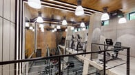 This two-level fitness facility delivers a comprehensive workout environment defined by its bold industrial aesthetic and high-end equipment. The architecture blends exposed concrete columns and walls with warm wood-paneled ceilings and a striking decorative feature wall adorned with an abstract mosaic pattern, creating a visually dynamic atmosphere throughout.

The main floor serves as the primary strength and conditioning area, featuring rubber flooring and an extensive array of equipment including power racks, barbells, weight plates, medicine balls, TRX suspension trainers, battle ropes, a heavy bag, and Concept2 rowing machines. Mirrored walls expand the sense of space, while exposed ductwork and pendant lighting contribute to the facility's industrial character. Large windows along the upper perimeter allow natural light to filter in, connecting the interior to the surrounding greenery outside.

A staircase with sleek cable-rail black metal railings leads to the upper mezzanine level, where cardio machines—including ellipticals and upright bikes—are arranged along the open railing, offering an elevated view of the main floor below.

A separate, enclosed cycling studio houses a row of indoor cycling bikes on light hardwood flooring, with recessed lighting and mirrored walls creating an intimate yet energizing environment. Access corridors finished with brushed metal accents and warm wood flooring complete this polished, purpose-built fitness destination.