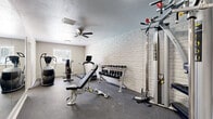 This single-floor fitness and recreation space is thoughtfully designed to support a well-rounded active lifestyle. The main workout area features white-painted brick walls that lend a clean, textured aesthetic, complemented by a textured ceiling and a ceiling fan that promotes air circulation during exercise. The durable, dark rubber-style flooring provides a practical and resilient surface throughout. The room is equipped with an impressive array of fitness equipment, including a treadmill with an integrated display, an elliptical trainer, an air resistance bike, a multi-station cable machine, an adjustable weight bench, and a tiered dumbbell rack stocked with a range of free weights. A full-length wall mirror in one section of the workout area enhances the sense of space and supports proper form during training. Natural light filters in through a window, adding brightness to the environment. An adjoining room, accessible through a doorway, transitions into a game room featuring hardwood-style flooring, a full-size pool table, and its own ceiling fan — making this space equally suited for leisure and entertainment. Together, these two connected rooms create a versatile and functional recreation area.