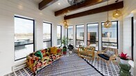 This charming sunroom blends modern farmhouse aesthetics with eclectic, inviting design details. The space is defined by crisp white shiplap walls that run the full perimeter, contrasted beautifully by bold, dark-stained exposed wood ceiling beams that add warmth and architectural character overhead. The flooring features a striking geometric patterned tile in black and white, grounded by a layered woven area rug that anchors the seating area.

Natural light pours in through an array of black-framed windows that line the walls, offering views of an adjacent outdoor terrace and the surrounding landscape. A set of black-framed French doors provides seamless access to the exterior, further blurring the boundary between indoors and out.

The space includes a rustic wooden potting bench topped with a galvanized metal basin, accompanied by a lush collection of potted plants and tropical greenery that lend a fresh, garden-inspired atmosphere. Vintage-style gooseneck wall sconces provide warm ambient lighting, complementing the rattan pendant lights that hang from the beamed ceiling.

Seating includes a bold floral-print sofa with vibrant jewel-toned pillows alongside rattan accent chairs, creating a relaxed, bohemian lounge feel. A glass-top bar cart rounds out the space, making this sunroom equally suited for casual gatherings or quiet relaxation.