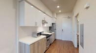This thoughtfully designed single-floor unit combines contemporary finishes with a clean, functional layout. The galley-style kitchen is a standout feature, outfitted with two-tone cabinetry — crisp white upper cabinets paired with light wood-grain lower cabinets — all complemented by white quartz countertops. Stainless steel appliances, including a gas range, dishwasher, and refrigerator, complete the well-appointed kitchen, while recessed lighting overhead keeps the space bright and inviting. Rich hardwood flooring flows consistently throughout the entire unit, lending warmth and cohesion to every room. The open living area is generously proportioned, featuring a built-in sliding-door closet and direct sightlines to both the kitchen and entry. A dedicated bedroom benefits from a large window that frames outdoor greenery and fills the room with natural light, creating a calm and airy atmosphere. The bathroom is finished with elegant white subway tile in a frameless glass-enclosed walk-in shower, accented with neutral tile trim details and chrome fixtures, adding a polished, spa-like quality. Recessed lighting and clean white walls throughout the unit reinforce a modern, minimalist aesthetic that is both practical and refined.