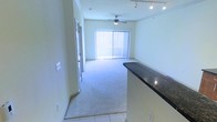This single-floor space features an open-concept layout that seamlessly connects the main living area to a kitchen counter space. The living room is finished with plush wall-to-wall carpeting, contributing to a comfortable and inviting atmosphere, while tiled flooring marks the transition near the entryway and kitchen zone.

A ceiling fan is centrally mounted overhead, providing both practical air circulation and a subtle decorative element. Track lighting along the ceiling offers additional illumination options, complementing the abundant natural light that streams in through a large sliding glass door fitted with vertical blinds. This door likely leads to a balcony or patio, enhancing the sense of openness and connectivity to the outdoors.

The kitchen area is partially visible and features a raised breakfast bar with a dark granite countertop, providing a functional divide between the cooking and living spaces. The walls throughout are finished in a neutral, light tone, keeping the interior feeling bright and airy. Crown molding adds a refined architectural detail, elevating the overall aesthetic of the space.