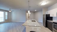 This two-bedroom, two-bathroom residence is thoughtfully designed on a single floor, offering a cohesive and functional layout. The open-concept living and kitchen area serves as the heart of the home, featuring light-toned luxury vinyl plank flooring that extends throughout, creating a clean and contemporary aesthetic.

The kitchen is a standout feature, centered around a large island with a striking granite countertop and an integrated sink. Pendant lighting hangs above the island, providing both task lighting and a stylish focal point. A stainless steel refrigerator is visible nearby, complementing the modern finishes. Recessed lighting and flush-mount fixtures illuminate the space efficiently.

The open layout connects the kitchen seamlessly to the living area, which offers ample room for furnishings and everyday use. Neutral gray walls and white trim provide a versatile backdrop throughout. Hallways lead to the two bedrooms and bathrooms, with a glimpse of a well-appointed bathroom visible in the background. Ceiling vents indicate a central HVAC system for year-round comfort. Overall, the space presents a bright, move-in-ready environment with clean, modern details.