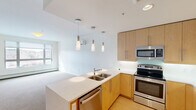 Located at 80 Winooski Falls Way, Unit 508, this two-bedroom, one-bathroom residence offers a thoughtfully designed single-floor layout with a clean, contemporary aesthetic throughout. The open-concept kitchen is a standout feature, showcasing light wood cabinetry, stainless steel appliances including a refrigerator, range, microwave, and dishwasher, along with a light stone countertop peninsula with a double sink. Stylish pendant lights hang above the peninsula, creating a warm and functional workspace that flows seamlessly into the carpeted living area. Natural light enters through windows that frame views of the Vermont landscape, adding a scenic backdrop to the comfortable living space. The primary bedroom is generously sized and features a large window with outdoor views, neutral carpet flooring, and baseboard heating. The second bedroom offers similar finishes with access to the hallway. A dedicated closet provides additional storage. The full bathroom is finished with large-format dark tile on both the floor and tub surround, a soaking tub, and clean white fixtures. Light wood flooring in the kitchen and entryway complements the neutral tones found throughout, creating a cohesive and modern living environment.