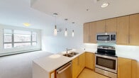 Located at 80 Winooski Falls Way, Unit 508, this two-bedroom, one-bathroom residence offers a thoughtfully designed single-floor layout with a clean, contemporary aesthetic throughout. The open-concept kitchen is a standout feature, showcasing light wood cabinetry, stainless steel appliances including a refrigerator, range, microwave, and dishwasher, along with a light stone countertop peninsula with a double sink. Stylish pendant lights hang above the peninsula, creating a warm and functional workspace that flows seamlessly into the carpeted living area. Natural light enters through windows that frame views of the Vermont landscape, adding a scenic backdrop to the comfortable living space. The primary bedroom is generously sized and features a large window with outdoor views, neutral carpet flooring, and baseboard heating. The second bedroom offers similar finishes with access to the hallway. A dedicated closet provides additional storage. The full bathroom is finished with large-format dark tile on both the floor and tub surround, a soaking tub, and clean white fixtures. Light wood flooring in the kitchen and entryway complements the neutral tones found throughout, creating a cohesive and modern living environment.