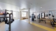 This fitness center is designed to support a comprehensive workout experience within a single-floor layout. The space features a light-filled interior, with natural light streaming in through multiple windows that brighten the clean, white walls and textured ceilings. Structural columns divide the floor plan into functional workout zones without compromising the open, airy feel of the space.

The gym is outfitted with a diverse selection of equipment to accommodate a variety of fitness routines. Strength training stations include cable and pulley machines, a weight bench, and free weights, while cardio options such as a rowing machine and stationary bike offer additional versatility. Large mirrors line one wall, enhancing the sense of depth and allowing users to monitor their form during exercises.

The flooring features a neutral gray surface with yellow accent striping that delineates different workout areas, contributing both to safety and visual organization. Recessed and surface-mounted lighting supplements the natural light to ensure the space remains well-illuminated throughout. The overall ambiance is clean, functional, and professional, reflecting a thoughtfully designed environment suited to meet a wide range of fitness needs.