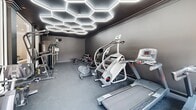 Located at 9400 Havenbrooke Way in Knoxville, Tennessee, this well-appointed fitness center delivers a contemporary workout environment with a distinctly modern aesthetic. The space is defined by its striking dark charcoal walls and matching flooring, creating a focused, high-energy atmosphere suited for serious exercise.

The standout design feature is the eye-catching honeycomb-patterned LED lighting system that spans the ceiling, casting bright, even illumination throughout the room while adding a bold architectural element. A large floor-to-ceiling mirror along one wall visually expands the space and supports form-checking during workouts.

Natural light enters through windows and glass-paneled double doors, which open to an outdoor area with a visible pool beyond — providing a refreshing visual connection to the exterior. The durable rubberized flooring throughout is practical and slip-resistant, ideal for high-intensity training.

Equipment is thoughtfully arranged across the room and includes multiple treadmills, an elliptical, a stair climber, a spin bike, and a rowing machine for cardio training. A dedicated strength zone features a multi-station cable machine, a dumbbell rack with a full weight selection, an adjustable bench, and additional resistance equipment. A wall-mounted TV screen rounds out the amenities, supporting interactive or on-demand workout sessions.