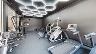 Located at 9400 Havenbrooke Way in Knoxville, Tennessee, this well-appointed fitness center delivers a contemporary workout environment with a distinctly modern aesthetic. The space is defined by its striking dark charcoal walls and matching flooring, creating a focused, high-energy atmosphere suited for serious exercise.

The standout design feature is the eye-catching honeycomb-patterned LED lighting system that spans the ceiling, casting bright, even illumination throughout the room while adding a bold architectural element. A large floor-to-ceiling mirror along one wall visually expands the space and supports form-checking during workouts.

Natural light enters through windows and glass-paneled double doors, which open to an outdoor area with a visible pool beyond — providing a refreshing visual connection to the exterior. The durable rubberized flooring throughout is practical and slip-resistant, ideal for high-intensity training.

Equipment is thoughtfully arranged across the room and includes multiple treadmills, an elliptical, a stair climber, a spin bike, and a rowing machine for cardio training. A dedicated strength zone features a multi-station cable machine, a dumbbell rack with a full weight selection, an adjustable bench, and additional resistance equipment. A wall-mounted TV screen rounds out the amenities, supporting interactive or on-demand workout sessions.