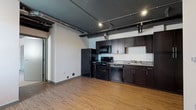 This single-floor loft-style unit showcases a distinctive industrial design, highlighted by dramatic dark-painted exposed ceilings with visible ductwork, beams, and track lighting throughout the main living and kitchen areas. The open-concept kitchen is well-appointed with rich dark espresso cabinetry, a full suite of black appliances including a refrigerator, electric range, built-in microwave, and dishwasher, complemented by light countertops and a stainless steel sink. Warm hardwood-style flooring extends through the kitchen and living areas, adding a welcoming contrast to the bold ceiling treatment above. The bedrooms offer plush carpeting underfoot, neutral white walls, and individual wall-unit HVAC systems, with windows that bring in natural light. The primary bedroom features direct access to a closet and an en-suite bathroom equipped with a corner neo-angle shower and wood-look flooring. A second bathroom serves the unit with a dark-cabinetry vanity, mirror, and wood-look flooring. The overall layout, as seen in the 3D floorplan, presents a well-organized and elongated space with a cohesive, contemporary urban character throughout.