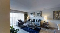 This single-floor space showcases a well-appointed living area designed with a contemporary aesthetic. The living room serves as the centerpiece, featuring a large sofa adorned with navy and grey accent pillows, paired with a matching upholstered armchair that contributes to a cohesive, sophisticated look. A deep navy shag area rug anchors the seating arrangement, adding texture and warmth to the light wood-toned flooring beneath.

A sleek rectangular coffee table with a glass or mirrored surface sits at the heart of the seating area, styled with decorative accents including metallic objects and fresh florals. Flanking table lamps on either side of the room cast a warm ambient glow, complementing the natural light that filters in through large floor-to-ceiling vertical blinds along one wall, hinting at an adjacent outdoor area or balcony.

The walls are adorned with two framed botanical artworks, adding a refined, curated touch to the neutral grey walls. Lush indoor plants are positioned throughout, enhancing the space with organic vitality. The textured ceiling adds subtle architectural character, contributing to the overall polished and inviting ambiance of this thoughtfully designed living space.