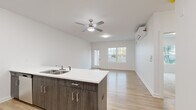 This well-designed single-floor residence in Troy, NY offers a thoughtful layout with one bedroom, two bathrooms, and a dedicated closet. The open-concept kitchen and living area serves as the heart of the home, where light wood-tone flooring flows seamlessly throughout. The kitchen is equipped with stainless steel appliances—including a refrigerator, range, and microwave—complemented by light cabinetry and a peninsula featuring dark wood-grain cabinetry below and a clean white countertop above, complete with a double-basin sink. A ceiling fan and wall-mounted mini-split unit provide year-round climate control in the living area, which benefits from natural light through a window and door leading to an outdoor deck surrounded by greenery. The bedroom is finished with plush carpeting and features a large window framing outdoor views of the surrounding neighborhood. It is serviced by a walk-in closet outfitted with wire shelving systems, offering ample storage. The bathrooms include a clean, all-white shower stall with chrome fixtures. Throughout, the space maintains a bright, modern aesthetic with white walls and crisp trim, creating a fresh and functional living environment.