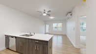 This well-designed single-floor residence in Troy, NY offers a thoughtful layout with one bedroom, two bathrooms, and a dedicated closet. The open-concept kitchen and living area serves as the heart of the home, where light wood-tone flooring flows seamlessly throughout. The kitchen is equipped with stainless steel appliances—including a refrigerator, range, and microwave—complemented by light cabinetry and a peninsula featuring dark wood-grain cabinetry below and a clean white countertop above, complete with a double-basin sink. A ceiling fan and wall-mounted mini-split unit provide year-round climate control in the living area, which benefits from natural light through a window and door leading to an outdoor deck surrounded by greenery. The bedroom is finished with plush carpeting and features a large window framing outdoor views of the surrounding neighborhood. It is serviced by a walk-in closet outfitted with wire shelving systems, offering ample storage. The bathrooms include a clean, all-white shower stall with chrome fixtures. Throughout, the space maintains a bright, modern aesthetic with white walls and crisp trim, creating a fresh and functional living environment.