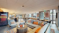 This single-floor communal amenity space is designed to foster both relaxation and productivity in a lively, open-plan layout. The interior is defined by a bold color palette anchored by striking burnt-orange accent walls, which contrast beautifully against light tile flooring, exposed concrete columns, and a crisp white drop ceiling with recessed and pendant lighting.

The lounge area features generous sectional and accent sofas adorned with colorful patterned throw pillows, arranged around organic wood coffee tables atop a vibrant area rug — creating an inviting space for casual gathering. Adjacent to the lounge, a fully equipped kitchen island with a white countertop and orange bar stools serves as a social hub, complete with stainless steel appliances, a range with hood, refrigerator, and open shelving illuminated by large dome pendant lights.

Multiple dining zones offer flexible seating with high-top bar tables and standard dining tables paired with warm wood chairs, accommodating both individual work and group interaction. Large floor-to-ceiling windows line the perimeter, flooding the space with abundant natural light and offering open views to the surrounding exterior. A single-cup coffee station adds a convenient amenity touch. The overall ambiance is energetic yet comfortable, blending modern design with functional, community-oriented living.