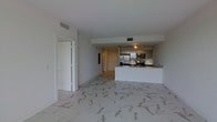 This single-floor space showcases a modern open-concept layout that seamlessly integrates the living area with the kitchen. The flooring throughout is a standout feature, comprised of large-format marble-style tiles with elegant grey veining set against a white background, lending a polished and sophisticated aesthetic to the entire space.

The kitchen is positioned along the rear wall and features a raised breakfast bar peninsula with a clean white finish, providing both a functional workspace and a natural divider between the kitchen and living area. Stainless steel appliances, including a refrigerator, range, and microwave, are integrated beneath dark cabinetry with under-cabinet track lighting, adding both practicality and a refined finish. A sink is conveniently located along the peninsula counter.

The living area is generously proportioned, benefiting from smooth white ceilings with recessed ventilation, contributing to a clean, uncluttered ambiance. A doorway leading to an additional room is visible, suggesting further functional space beyond the open living area. The overall design palette of whites and neutrals creates a bright, airy environment throughout.