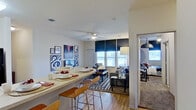 This single-floor residence offers a thoughtfully designed open-concept layout that seamlessly connects the kitchen, dining, and living areas. The kitchen is outfitted with rich dark-wood cabinetry, black appliances including a range and dishwasher, a double sink, and generous counter space. A breakfast bar with bar-height seating extends from the kitchen, providing a casual dining option that flows naturally into the main living space. The living room is bright and welcoming, featuring hardwood-style flooring, a ceiling fan, and multiple windows dressed with navy roller shades that allow for natural light while maintaining privacy. Two distinct bedrooms each carry their own character — one features a dramatic tiled accent wall, a statement pendant light, and a window with a roller shade, while the other showcases a crystal chandelier and dark curtain panels for an elegant touch. Each bedroom benefits from dedicated closet space with built-in shelving and hanging storage. The bathrooms are finished with dark-wood vanities, round mirrors, and shower-tub combinations. Completing the unit is a dedicated in-unit laundry room with a full-size washer and dryer and overhead shelving for added storage convenience.