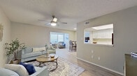 This single-story unit offers a thoughtfully organized layout across a comfortable footprint. The open-concept living area is anchored by light wood-style flooring and neutral warm-gray walls, creating a cohesive and inviting atmosphere. A ceiling fan with integrated lighting adds both function and visual interest to the main living space, which flows seamlessly into a secondary sitting area bathed in natural light from large windows fitted with blinds.

The kitchen features white shaker-style cabinetry, white quartz-style countertops, a subway tile backsplash, and a full suite of black appliances including a refrigerator, range, microwave, and dishwasher. A pass-through window connects the kitchen to the living area, enhancing the open feel. Adjacent to the kitchen, a defined dining area accommodates a bistro-height table and chairs.

The bedroom is a comfortable, private retreat with direct access to a walk-in closet equipped with multi-tier shelving. The bathroom includes a tub-shower combination with white tile surround, a dark-framed vanity mirror, and a single-sink countertop with modern fixtures. Light wood-style flooring continues throughout all rooms.