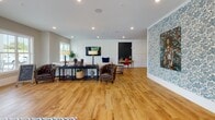 Located at 300–384 N Liberty St in Spartanburg, South Carolina, this single-floor community space combines modern design with welcoming functionality. The layout flows seamlessly across an open floor plan, unified by warm hardwood flooring that runs throughout the entire space.

The main lounge area is generously proportioned, featuring a paneled accent wall with a wall-mounted flat-screen TV, tufted leather armchairs, and a sectional sofa arranged around a central coffee table. A striking floral wallpapered wall adds an artistic focal point, while recessed lighting provides consistent illumination across the space. Large windows along the perimeter allow ample natural light to fill the room, with direct views of the outdoor deck and surrounding greenery visible through full-light glass doors.

A dedicated kitchen and bar area anchors one side of the space, finished with white shaker cabinetry, a white subway tile backsplash, and a sleek white countertop island with leather-topped barstools. Industrial-style pendant lights hang overhead, and a mounted television and coffee station make this area ideal for casual gatherings. Decorative blue ceramic plates line the backsplash, adding a curated, homey touch.

Overall, the space presents a clean, contemporary aesthetic with thoughtful furnishings, eclectic décor accents, and a bright, open ambiance suited for socializing and community engagement.
