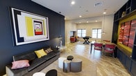 This stylish single-floor community lounge space is thoughtfully designed to encourage social interaction and leisure. The interior makes a bold visual statement with a striking dark navy accent wall adorned with a large-scale, colorful geometric artwork that anchors the main seating area. A plush charcoal sofa with yellow and patterned throw pillows offers a comfortable focal point, complemented by a cluster of modern cylindrical side tables in varying metallic and neutral finishes.

The open floor plan flows seamlessly into distinct activity zones. A game area features a foosball table, while a set of high-top tables and red accent chairs provide a casual dining or gathering spot. A pair of red lounge chairs near a fireplace creates an inviting nook for more intimate conversation. Sleek pendant lighting and recessed fixtures illuminate the space throughout, enhancing its contemporary aesthetic.

Light hardwood-style flooring runs through the entire area, adding warmth and cohesion. Natural light filters in through multiple windows, contributing to the bright and energetic ambiance. The overall design palette — combining deep navy, bold red, yellow, and neutral tones — gives this communal space a dynamic, modern character that is both functional and visually engaging.
