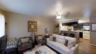 This single-floor, 2-bedroom, 2-bathroom residence offers a thoughtfully designed layout that balances comfort and functionality. The living area serves as the heart of the home, featuring warm neutral-toned walls, a textured ceiling, and a ceiling fan with integrated lighting that enhances the welcoming ambiance. The space is furnished with a plush white sofa accented by decorative pillows, complemented by a pair of patterned armchairs arranged around a glass-top coffee table atop a soft area rug. Decorative wall art in ornate gold frames adds a classic touch, while a mounted deer head display contributes a distinctive, rustic accent. The living area opens seamlessly into a dining and kitchen area, visible in the background, where bar-height seating and overhead cabinetry suggest a practical and connected entertaining space. Dark hardwood-style flooring grounds the interior, providing contrast to the light-colored furnishings. With its open flow between living, dining, and kitchen areas, this residence is well-suited for both everyday living and hosting guests. The cohesive design palette of warm neutrals and gold accents lends the space a polished, inviting character throughout.