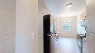 This single-floor unit showcases a clean, contemporary design highlighted by light wood-tone luxury vinyl plank flooring that runs consistently throughout the living spaces. The kitchen is a standout feature, designed in a galley-style layout with light wood cabinetry, white quartz countertops, and black appliances including an electric range, over-the-range microwave, and refrigerator. A peninsula with an integrated sink opens the kitchen toward the adjacent living and dining area, where a decorative chandelier and a large window bring in natural light. A wall-unit HVAC system, a secondary window, and a glass-panel exterior door provide both comfort and outdoor access in the main living area. The bedrooms are generously proportioned, finished with neutral tones and closet storage, with windows offering outdoor views. The bathroom features patterned tile flooring with a classic geometric motif, a wood-toned vanity with dark hardware, a tub-shower combination, and built-in shelving for storage. Throughout the unit, neutral wall tones, clean white trim, and consistent modern fixtures contribute to a cohesive and move-in-ready aesthetic.