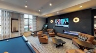 Located along Berewick Commons Parkway in Charlotte, NC, this single-floor clubhouse amenity space is thoughtfully designed with a modern, upscale aesthetic throughout. The grand entry lobby sets an impressive tone, featuring a striking crystal chandelier, herringbone-patterned tile flooring, and dark, textured wall paneling that frames the corridor leading toward large glass entry doors.

The social lounge area offers a welcoming gathering space outfitted with round tables and a mix of neutral and rust-colored accent chairs arranged around a large wall-mounted flat-screen display flanked by built-in shelving with curated décor and warm pendant lighting overhead.

A dedicated leasing or business office provides a more intimate and professional environment, highlighted by rich textured wallcovering, elegant circular gold wall sconces, a sleek central work table, and built-in shelving with decorative objects. A barn-style sliding door adds a contemporary architectural detail.

The game and entertainment room rounds out the space with a billiards table, leather lounge chairs, a linear fireplace, and a large wall-mounted television. Oversized windows dressed with patterned drapery flood the room with natural light and offer views of the surrounding community. Fitness equipment is also present, adding a wellness component to this versatile amenity space.