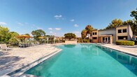 Located at 1735 Brantley Rd in Fort Myers, Florida, this outdoor community amenity space centers around a generously sized swimming pool with clearly marked depth gradients ranging from a shallow entry point to a deep end. The pool features a classic rectangular design with step entries, metal handrails, and a clean paver deck that surrounds the perimeter, providing ample space for lounging and relaxation.

Adjacent to the pool, a white pergola structure offers a shaded seating area furnished with chairs and tables, making it an ideal spot to unwind out of the Florida sun. Vibrant orange umbrellas add a pop of color to the outdoor seating zones scattered throughout the deck area. The space is framed by mature tropical trees, including towering palms and broad canopy trees, along with lush, colorful shrubs that create a verdant, resort-like atmosphere.

The surrounding buildings feature a combination of stone and siding exteriors with large windows, complementing the overall aesthetic of the community. A dedicated garden area with mature shade trees and ornamental plantings adds a natural, tranquil dimension to the space. The entire area benefits from abundant Florida sunshine, with an open sky view that enhances the bright, welcoming ambiance of this well-maintained outdoor retreat.