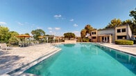 Located at 1735 Brantley Rd in Fort Myers, Florida, this outdoor community amenity space centers around a generously sized swimming pool with clearly marked depth gradients ranging from a shallow entry point to a deep end. The pool features a classic rectangular design with step entries, metal handrails, and a clean paver deck that surrounds the perimeter, providing ample space for lounging and relaxation.

Adjacent to the pool, a white pergola structure offers a shaded seating area furnished with chairs and tables, making it an ideal spot to unwind out of the Florida sun. Vibrant orange umbrellas add a pop of color to the outdoor seating zones scattered throughout the deck area. The space is framed by mature tropical trees, including towering palms and broad canopy trees, along with lush, colorful shrubs that create a verdant, resort-like atmosphere.

The surrounding buildings feature a combination of stone and siding exteriors with large windows, complementing the overall aesthetic of the community. A dedicated garden area with mature shade trees and ornamental plantings adds a natural, tranquil dimension to the space. The entire area benefits from abundant Florida sunshine, with an open sky view that enhances the bright, welcoming ambiance of this well-maintained outdoor retreat.