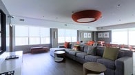 This single-floor space showcases a contemporary open-plan design centered around a spacious living area. The room is anchored by a large, sectional sofa in a neutral gray upholstery, accented with vibrant orange and green throw pillows that add bold pops of color throughout. A striking burnt-orange drum pendant light serves as a dramatic focal point on the white ceiling, lending warmth and a sense of architectural intention to the space.

The living area features smooth hardwood-style flooring in a warm tone, complemented by sleek, sculptural coffee tables with curved white bases. A low bench with red legs near the windows adds a functional yet decorative touch. Floor-to-ceiling roller shades line the expansive windows along the perimeter walls, diffusing natural light while maintaining views of the surrounding urban skyline.

Transitioning toward the rear of the space, a dining area is visible, furnished with orange upholstered seating and a glass-top table, maintaining the cohesive color palette. Framed artwork on the gray-toned walls adds subtle visual interest. The overall ambiance is modern, vibrant, and urban, with a carefully curated design that balances bold accents against neutral architectural elements.