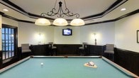 Located at 1455 Washington Blvd in Stamford, Connecticut, this single-floor recreational space is designed for leisure and socializing. The centerpiece of the room is a full-size billiards table, perfectly positioned beneath an ornate wrought-iron chandelier fitted with three large drum pendant shades that cast a warm, focused light over the playing surface. The room features a classic aesthetic, with dark wood wainscoting and trim accenting the crisp white upper walls, adding a sophisticated, club-like ambiance. A wall-mounted flat-screen television is centrally placed, ensuring visibility from throughout the room. Flanking the TV are two bar-height table-and-stool seating areas, each illuminated by elegant wall sconces, providing comfortable spots for spectators or casual conversation. The ceiling features a subtle tray detail with dark crown molding that reinforces the refined character of the space. The overall design balances a traditional, upscale aesthetic with functional amenities, making it well-suited as a social and entertainment hub.