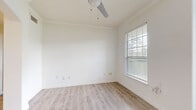 This efficiently designed single-story unit offers a thoughtful layout across its compact footprint, accommodating a kitchen, bedroom, bathroom, walk-in closet, and hallway. The living and dining areas feature light-toned wood-look flooring and crisp white walls accented by crown molding, with multiple windows fitted with horizontal blinds allowing generous natural light and outdoor views. An arched passageway connects the living space to the kitchen, which is outfitted with white raised-panel cabinetry, light quartz countertops, a marble-style tile backsplash, and a full suite of stainless steel appliances including a range, over-the-range microwave, and refrigerator. The bedroom is finished with neutral carpet, a ceiling fan, and windows that further enhance the bright, airy atmosphere. An adjoining walk-in closet provides practical storage with white wire shelving systems lining the walls and a central shelving tower. The bathroom features a white vanity with ample cabinet storage, a framed mirror, lighted vanity fixture, and wood-look tile flooring. Throughout, the unit maintains a clean, neutral palette that creates a cohesive and functional living environment.