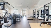 Located at 23615 Kingsland Blvd in Houston, TX, this fitness center offers a generously sized, single-floor exercise space designed to support a full range of workout routines. The facility is defined by its striking vaulted ceilings, which lend a sense of openness and volume throughout the main floor area. Large, multi-pane windows line the walls, inviting abundant natural light and providing views of the surrounding exterior — creating an energizing atmosphere for exercise.

The exercise room is outfitted with a comprehensive selection of equipment. A dedicated cardio zone features treadmills, ellipticals, and stair climbers positioned along the windowed walls, taking full advantage of the natural light. The strength training area includes power racks with bench press setups, plate-loaded barbells, and a variety of selectorized resistance machines, catering to both free-weight and machine-based training. A large wall mirror expands the sense of space and supports proper form during workouts.

The flooring transitions between durable rubber fitness flooring in the workout zones and hard-surface flooring near the entrance, which is framed by an elegant arched doorway and glass French doors. Ceiling fans and linear LED lighting ensure the space remains comfortable and well-lit. The clean, modern, white interior palette gives the facility a bright and professional aesthetic throughout.
