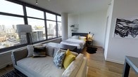 This thoughtfully designed studio occupies an elevated position within a high-rise building, offering a seamlessly integrated living and sleeping space with a clean, contemporary aesthetic. The most striking feature is the expansive floor-to-ceiling window wall that floods the interior with natural light and frames an impressive panoramic view of the surrounding urban skyline — a dramatic backdrop that defines the ambiance of the entire space.

The open-plan layout is both functional and inviting. The living area is anchored by a plush, neutral-toned sofa accented with geometric and boldly colored throw pillows in shades of yellow and grey, adding a vibrant, modern touch. A low-profile coffee table serves as a central focal point between the seating and sleeping zones. The sleeping area features a well-appointed bed with coordinated linens, complemented by a sleek metal-framed nightstand and wall-mounted shelving for added storage. A stylish arc lamp provides targeted task lighting.

Warm hardwood flooring runs throughout, grounding the space and adding a natural contrast to the cool white walls and dark window frames. The overall atmosphere is bright, urban, and sophisticated — ideal for those seeking a polished, contemporary living experience in the heart of the city.