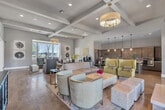 Located in Murrieta, CA, this single-floor community clubhouse offers a well-appointed, open-concept layout that seamlessly integrates a spacious living area with a fully equipped kitchen. The living room features soaring ceilings with decorative coffered and exposed beam detailing, creating a sense of grandeur and openness. Warm hardwood flooring flows throughout, complementing the neutral color palette and contemporary furnishings, including upholstered lounge chairs, a natural wood drum coffee table, and a tufted bench. Large bay windows invite abundant natural light, offering views of the surrounding outdoor landscape. A fireplace anchors one end of the living area, accompanied by a mounted TV and a patterned accent wall, while a coffee station adds convenience for guests. The kitchen is a standout feature, showcasing grey shaker-style cabinetry, a mosaic tile backsplash, granite countertops, stainless steel appliances, and a large center island with bar seating beneath a row of pendant lights. Curated wall art and tasteful décor accents contribute to the polished, welcoming ambiance, making this space well-suited for social gatherings and community events.