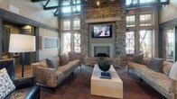 This thoughtfully designed single-story space in Nashville, Tennessee, showcases a compelling fusion of rustic craftsmanship and contemporary elegance. The architecture is defined by soaring vaulted ceilings with exposed dark timber beam trusses that draw the eye upward, creating a dramatic sense of volume throughout the interior. Wide-plank hardwood flooring grounds the space, complementing the warm, neutral tones of the wood-paneled walls.

A striking floor-to-ceiling stone fireplace serves as the focal point of the lounge area, flanked by large multi-pane windows that flood the space with abundant natural light and offer scenic views of the surrounding Tennessee landscape. Plush sofas and upholstered armchairs are arranged to encourage comfortable conversation, while a wall-mounted television and a chalkboard panel add functional touches to this inviting gathering area.

The dining area transitions seamlessly from the lounge, featuring marble-topped tables, sculptural white chairs, and built-in banquette seating set within a nook defined by dark wood posts. Pendant lighting in brushed brass and matte black accents adds warmth overhead. A glass-partitioned workspace and service area provide additional functionality. Throughout, teal-accented barn-style doors and curated artwork contribute personality, while expansive windows ensure the outdoors remain a constant, breathtaking presence.