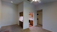 This single-floor residence offers a thoughtfully arranged open layout that maximizes functionality and flow. The main living area features notably tall ceilings equipped with a ceiling fan, creating an airy and spacious atmosphere. Neutral carpet flooring runs throughout the living space, contributing to a clean and cohesive aesthetic.

A pass-through counter with dark wood cabinetry connects the living area to the kitchen, providing a convenient serving and workspace. The kitchen itself is well-appointed with warm wood cabinetry, a white electric range and oven, a dishwasher, and ample counter space, all set on hard-surface flooring that is practical for a cooking environment.

Adjacent to the kitchen, a dedicated laundry area houses a stacked washer and dryer unit behind a set of bifold doors, keeping utilities neatly concealed. A closet nearby offers additional storage.

The overall color palette throughout the home is light and neutral, with white walls that reflect natural and ambient light effectively. The ceiling-mounted ventilation and well-placed lighting fixtures add to the practical design of the space. The layout is efficient yet open, making it easy to navigate between the living, cooking, and utility areas seamlessly.