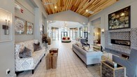 This sophisticated single-floor amenity space is designed to impress, offering a seamless blend of upscale design and functional comfort across several distinct gathering areas. The grand entrance lobby features soaring vaulted ceilings clad in warm natural wood planking, tile flooring, and an abundance of natural light streaming through large arched and floor-to-ceiling windows that frame views of a resort-style pool courtyard beyond.

The main lounge area is anchored by a decorative fireplace with an intricate tiled surround, a wall-mounted television, and a coffered ceiling with ambient lighting that adds warmth and depth. Plush sectional seating and accent chairs are arranged to encourage conversation and relaxation. Adjacent seating areas continue the refined aesthetic with tufted upholstery, mirrored accents, and carefully curated décor.

A dedicated game and entertainment room features dual wall-mounted screens above a stylish bar counter with barstool seating, pendant lighting with a Moorish-inspired tile backdrop, and a full billiards table — making it an ideal space for social gatherings. Intimate dining and lounge nooks positioned near the large glazed windows offer picturesque outdoor views, completing this thoughtfully appointed community space.