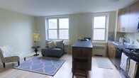 This single-floor space offers a clean, functional layout filled with natural light streaming through multiple large double-hung windows that frame views of surrounding urban architecture and greenery. The interior is finished with light hardwood flooring throughout, lending a warm and airy quality to the space. Walls are painted in a soft, neutral tone that complements the natural light and enhances the sense of openness.

The living area is anchored by a plush gray chaise lounge set atop a patterned blue area rug, creating a cozy yet stylish seating nook. A decorative vase with yellow blossoms on a small side table adds a touch of color and life to the corner. Radiator units beneath the windows indicate a classic heating system well-suited for urban living.

Adjacent to the living area, a dark-stained wooden counter or bar unit with a black top and accompanying stool serves as a natural divider, suggesting a transitional dining or workspace zone. A second geometric area rug nearby reinforces the delineation of functional areas within the open floor plan. The overall ambiance is calm, practical, and well-lit.