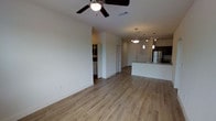 This single-floor space is designed with an open-concept layout that seamlessly connects the living area to the kitchen, creating a bright and airy environment. The living area is finished with light-toned wood-style flooring that extends throughout, lending warmth and continuity to the space. A ceiling fan with an integrated light fixture is centrally mounted, providing both airflow and ambient lighting.

The kitchen is positioned toward the rear of the open floor plan and features a sleek, light-colored countertop island with an integrated sink. A stainless steel refrigerator is visible, complementing the modern aesthetic of the space. Overhead, a combination of pendant lights and track lighting illuminates the kitchen area, adding a contemporary touch.

The neutral white walls provide a clean backdrop throughout, while the open passageways between areas enhance the sense of spaciousness. Additional doorways lead to separate rooms, including what appears to be a utility or laundry area to one side, adding functional versatility. The overall ambiance is modern, clean, and well-lit, making the space feel open and move-in ready.